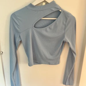 Blå långärmad crop top  - Trendig ljusblå långärmad topp med ribbad struktur och cool cut out-detalj vid axeln. Croppad modell med hög hals som ger en modern vibe. Perfekt för dig som vill sticka ut lite extra.