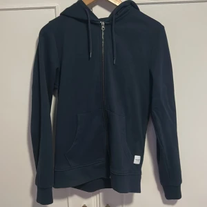 Mörkblå hoodie från Jack & Jones XS - Mörkblå hoodie från Jack & Jones Essentials i storlek XS. Tröjan har dragkedja framtill, huva med snörning och två fickor. Perfekt för chill dagar och enkel att matcha med jeans eller joggers.