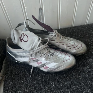 Adidas Predator  - Snygga Adidas Predator fotbollsskor i silver med vita och vinröda detaljer. ( bellinghams) Skorna har snörning, mönstrad ovandel och klassiska dobbar för gräsplan. Lätta och smidiga med cool design och logga på plösen. Perfekta för dig som vill sticka ut på planen.