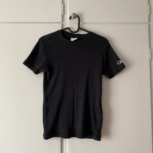 Svart t-shirt från Champion - Säljer en oanvänd svart ribbad t-shirt från Champion. Klassisk modell med korta ärmar och rund hals. Champion-logga broderad på ärmen. Skön och stretchig bomullskvalitet som sitter snyggt och passar till allt.