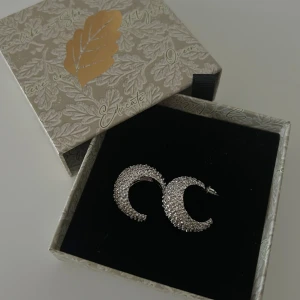 Glittriga silverörhängen  - Supersnygga örhängen i form av halvmånar, täckta med glittrande stenar. De är i silverfärg och har en cool, unik design som verkligen sticker ut. Fick som present, aldrig använda 