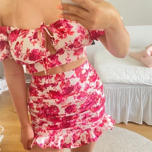 Blommig offshoulder sett - Superfint sett med rosa och röda blommor på vit botten. Offshoulder-modell med volangkant och snörning framtill. Settet har figurnära passform och volang nertill, perfekt för sommarens alla tillfällen.