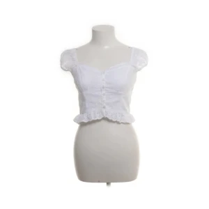 Vit blus med volang och knappar - Superfin vit blus i bomull med korta puffärmar, knappar framtill och volangkant nertill. Söt broderad spetsdetalj och croppad passform som ger en romantisk vibe. Perfekt för varma dagar och sommarhäng.