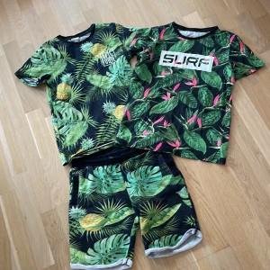 Tropisk t-shirt H&M x K. Kijak - Snygg t-shirt från H&M x K. Kijak i tropiskt mönster med gröna blad och gula ananaser på svart botten. Rund halsringning och korta ärmar. Perfekt för dig som gillar färgstarka prints och sommarkänsla.