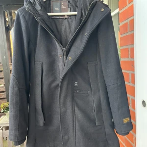 Svart kappa från G-Star RAW med huva - Svart kappa från G-Star RAW med snygg huva och dragkedja framtill. Jackan har flera fickor, diskreta logodetaljer och en stilren look. Tillverkad i ullblandning som håller dig varm under kyliga dagar. Perfekt för dig som gillar en clean och urban stil. Dock är vänster fickas dragkedja trasig. 