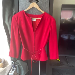 Röd kavaj med knyt från My time - Snygg röd kavaj från My time med trekvartsärm och smickrande passform. Jackan har ett smalt knytband i midjan och stilren, öppen halsringning. Perfekt för dig som vill sticka ut med färg och enkel design.
