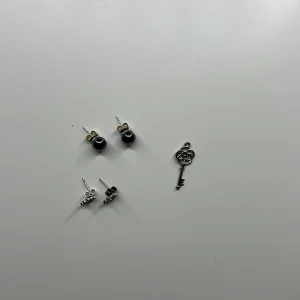 Två par örhängen och en nyckelberlock - Säljer två par örhängen: ett par med svarta glansiga kulor och ett par små silvriga örhängen med gnistrande stenar. Medföljer även en söt liten berlock i form av en silverfärgad nyckel. Perfekt för dig som gillar att mixa och matcha smycken.