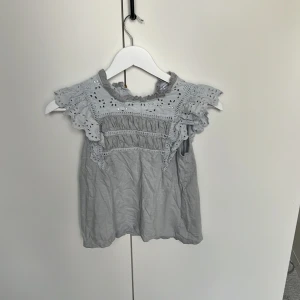 Ljusgrå blus med spets från Lindex - Supersöt ljusgrå blus från Holly & Whyte by Lindex i bomull. Blusen har broderade spetsdetaljer och volang vid axlarna, samt en högre halsringning med romantiska hålmönster. Perfekt för dig som gillar en feminin och trendig look.