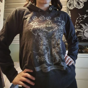Svart hoodie med tryck och huva - Cool svart hoodie med stort detaljerat tryck framtill i grått och silver, med texten 'Legend' och svärdsmotiv. Tröjan har huva med snörning och långa ärmar. Perfekt för dig som gillar en edgy och avslappnad stil. Huvan är väldigt liten så svår att använda.