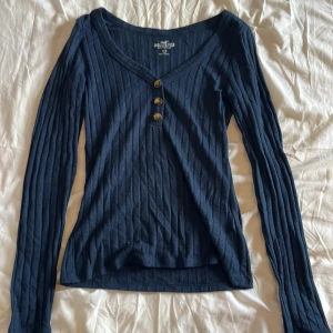 Mörkblå ribbad tröja Hollister - Supersnygg mörkblå ribbad tröja från Hollister med v-ringning och tre knappar framtill. Långärmad och i mjuk bomull, perfekt till jeans eller kjol. Sitter normal och har en klassisk men trendig vibe.