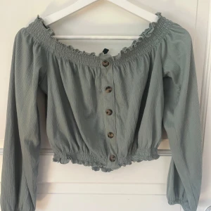 Grön offshoulder blus med knappar - Supersöt ljusgrön blus med offshoulder-modell och långa ärmar. Blusen har knappar framtill, smockad resår vid halsringning och midja samt volangdetaljer. Perfekt för dig som gillar romantisk och trendig stil.