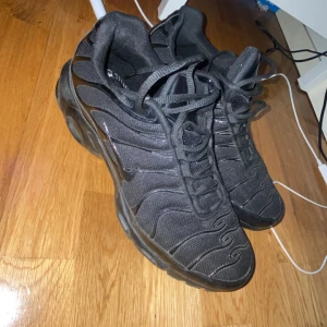 Svarta Nike Air Max Plus TN sneakers - Säljer ett par svarta Nike Air Max Plus TN sneakers med ikoniska vågiga detaljer och Tn Air-logga på hälen. Skorna har mesh och syntetmaterial, snörning och synlig Air Max-dämpning i sulan. Perfekta för dig som gillar streetstyle och chunky sneakers.