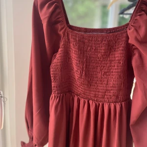 Vinröd långklänning - Vinröd långklänning med smockad överdel och ärmslut, puffiga långärmar och fyrkantig ringning. Klänningen har volang nertill och är i ett mjukt, flowy material. Oanvänd.