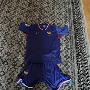 Frankrike Nike fotbollströja blå - Säljer en blå Frankrike fotbollströja från Nike med FFF-logga och broderad tupp på bröstet. Tröjan har korta ärmar, röda detaljer vid sidorna och vit Nike-logga. Materialet är lätt och andas, perfekt för match eller träning. Matchande shorts medföljer.
