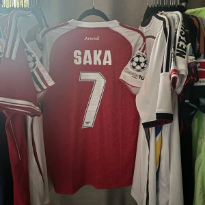 Arsenal Saka fotbollströja 2025/26 - Arsenal hemmatröja med Saka 7-tryck, Champions League-märke på ärmen och Emirates Fly Better sponsorlogga. Röd bas med vita detaljer och klassiska Adidas-ränder på axlarna. Tillverkad i lätt och ventilerande material, perfekt för match eller supporterliv.