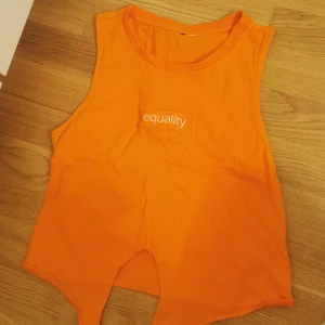 Orange croppad linnetopp med tryck - Cool orange ärmlös linnetopp med texten 'equality' broderad framtill. Toppen har croppad passform och spetsig nederkant framtill för en edgy look. Perfekt för dig som vill sticka ut med färg och statement.