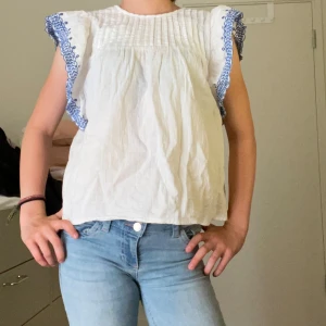 Vit blus med blå brodyr från MNG - Supersöt vit blus från MNG med volangärmar och blå broderade detaljer längs ärmsluten. Blusen har en luftig passform och är tillverkad i ett lätt bomullstyg, perfekt för varma dagar. Snyggt rynkad upptill och riktigt fräsch till jeans eller kjol.