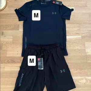 Under Armour set svart - Svart t-shirt från Under Armour med diskret logga på bröstet och tryckt text längs sidan. T-shirten är kortärmad och har en sportig, normal passform. Perfekt för dig som gillar att träna och vill ha en stilren look.