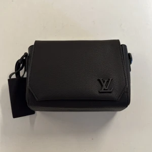 Svart axelväska från Louis Vuitton - Stilren svart axelväska från Louis Vuitton i exklusivt skinn. Väskan har ett klassiskt LV-emblem framtill, justerbar axelrem och praktisk dragkedjeficka på baksidan. Perfekt storlek för det viktigaste och en snygg accessoar till din outfit.