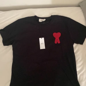 Svart t-shirt med rött hjärta från Ami - Svart t-shirt från Ami Alexandre Mattiussi med ikoniskt rött hjärta och A-brodyr på bröstet. Klassisk passform, rund hals och korta ärmar. Tillverkad i mjuk ekologisk bomull för en clean och stilren look.