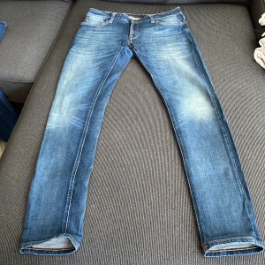 Blå skinny jeans från Nudie Jeans - Snygga blå skinny jeans från Nudie Jeans med slitningar och ljusa partier på benen. Klassisk femficksmodell med orangea sömmar och logga bak. Jeansen är i stretchig bomull och har en smal passform som sitter tajt längs benen. Små i storleken