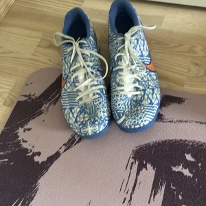 Nike CR7 Air Zoom blå/vita fotbollsskor - Säljer ett par Nike CR7 Air Zoom fotbollsskor med unikt blått och vitt mönster, orange swoosh och CR7-detaljer. Skorna har snörning, platt sula och är tillverkade i syntetmaterial. Perfekta för dig som vill sticka ut på planen!