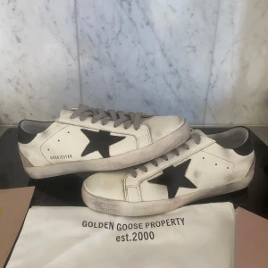 Golden Goose Superstar sneakers vit/svart - Golden Goose Superstar sneakers i vitt skinn med svart stjärna på sidan och svart häl. Grå snörning och beige mockadetalj på hälen. Klassisk rund tå och platt sula. Snyggt slitna detaljer för en cool streetstyle-look.