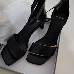 Svarta Vagabond sandaler med klack - Stilrena svarta sandaler från Vagabond med fyrkantig tå, bred rem över foten och justerbar rem runt vristen. Skorna har hög klack på ca 7 cm och är tillverkade i skinn. Perfekta för dig som gillar minimalistisk och modern look.