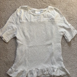 Vit blus med volang från Gina Tricot - Supersöt vit blus från Gina Tricot med smockad framsida och volangkant nedtill. Blusen har korta ärmar och rund halsringning. Tillverkad i mjuk bomull.