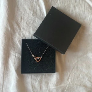 Halsband med hjärta i silver och roséguld - Fint halsband med tunn silverkedja och ett hängsmycke i form av två sammanlänkade hjärtan, ett i roséguld och ett i silver. Perfekt accessoar för att lyfta din look med en söt detalj. Aldrig använd och ingen aning var den kommer från men antagligen glitter!