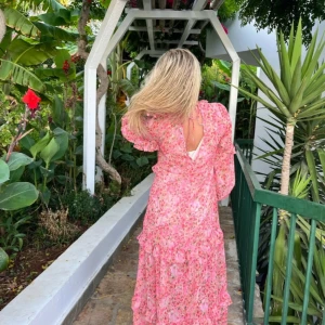 Rosa blommig långklänning med volanger - Superfin långklänning i rosa med blommigt mönster och volangdetaljer. Klänningen har långa ärmar, v-ringning och öppen rygg med knyt. Perfekt för sommarens alla tillfällen och ger en romantisk vibe.