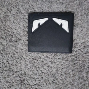 Svart plånbok med ögon - Säljer en svart plånbok från  i skinn med ikoniska vita monsterögon på framsidan. Plånboken har flera kortfack och ett fönster för ID. Perfekt för dig som vill ha en stilren men ändå unik accessoar.