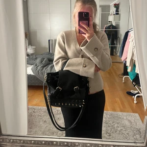 Svart axelväska med nitar och spikes - Cool svart axelväska i skinnimitation med justerbar rem och handtag. Väskan har guldiga spikes och nitar längs nederkanten, två spännen framtill och dragsko. Perfekt för dig som vill sticka ut med en edgy accessoar.