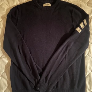 Mörkblå stickad tröja Zadig - Mörkblå stickad tröja från Zadig med rund hals och långa ärmar. På ena  ärmen finns tre breda vita ränder som ger en sportig touch. Tröjan är mjuk och passar perfekt till jeans eller chinos. Köptes från barnavdelningen! Storlek 16-M… frakt betalar köparen 