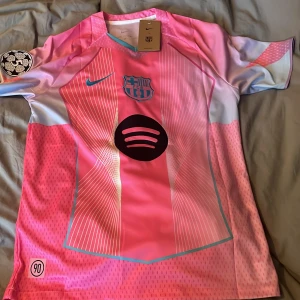 FC Barcelona Lamine Yamal Nike rosa tröja - Snygg FC Barcelona fotbollströja i rosa och ljusblått från Nike med Lamine Yamal och nummer 10 på ryggen. Tröjan har korta ärmar, tryckt Spotify-logga, klubbmärke och UEFA Foundation-märke på ärmen. Tillverkad i lätt och ventilerande material.