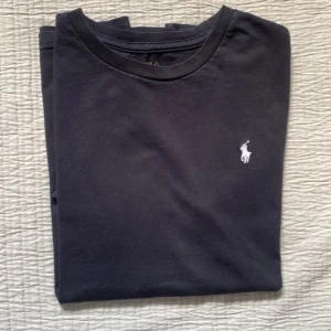 Svart t-shirt från Polo Ralph Lauren - Svart t-shirt från Polo Ralph Lauren med klassisk rund hals och vit broderad logga på bröstet. Tillverkad i mjuk bomull som känns skön mot huden. Perfekt basic-plagg med stilren design och normal passform.