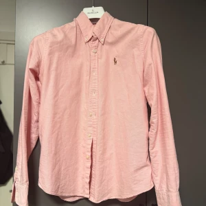 Rosa skjorta från Ralph Lauren - Snygg rosa skjorta från Ralph Lauren med klassisk button-down krage och broderad logga på bröstet. Skjortan har långa ärmar, knappar framtill och är tillverkad i mjuk bomull. Perfekt för dig som gillar stilrena och tidlösa plagg. Säljer den då den är lite liten för mig. Pris kan diskuteras vid snabb och smidig affär 🌸