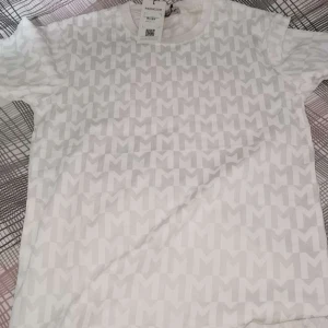 Vit Moncler t-shirt med mönster - Snygg vit t-shirt från Moncler med diskret heltryckt M-mönster över hela plagget. Klassisk rund halsringning och korta ärmar. Perfekt för dig som gillar stilrena och exklusiva märken. Materialet känns mjukt och skönt mot huden.