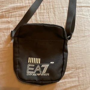 EA7 väska - Snygg svart axelväska från EA7 Emporio Armani med justerbart band och vit logga framtill. Väskan har dragkedja upptill, innerfack med dragkedja och ytterfack. Tillverkad i slitstark polyester, perfekt för att bära dina viktigaste grejer.