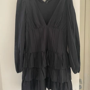 Svart volangklänning från Zara - Supersnygg svart kortklänning från Zara med djup v-ringning, långa ärmar och flera volanglager. Klänningen har en lätt randig struktur i tyget och ger en cool, trendig look. Perfekt för dig som gillar statement-plagg med mycket rörelse. Tyvärr lite sönder i nacken. 