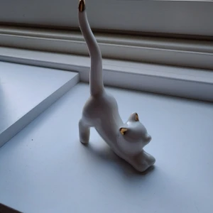 Vit kattformad ringhållare i keramik - Superfin ringhållare i form av en stretchande katt i vit keramik med detaljer i guld på öron och svanstipp. Perfekt att förvara dina ringar på svansen eller bara ha som söt inredningsdetalj på smyckesbordet.