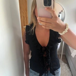 Svart ribbad topp med volang och snörning - Supersnygg svart ribbad topp med djup v-ringning, volangkant längs axlar och fram, samt snörning framtill. Ärmlös modell som är perfekt till jeans eller kjol. Ger en cool och trendig vibe.