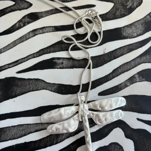 Silverfärgat halsband med trollslända - Unikt halsband i silverfärg med en stor trollslända som hänge. Kedjan är ormlänk och hänget har en cool, hamrad yta som ger extra karaktär. Perfekt accessoar för dig som gillar naturinspirerade smycken och vill sticka ut.