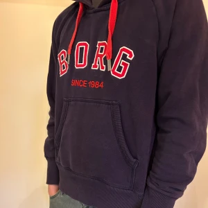Björn Borg hoodie - Snygg mörkblå hoodie från Björn Borg med röd text och röda snören. Stor känguruficka framtill och huva. Skön bomullsblandning och klassisk passform, perfekt för chill dagar eller när du vill ha en sportig look.
