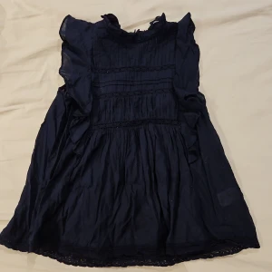 Mörkblå blus med volang från Lindex XS - Supersöt mörkblå blus från Holly & Whyte by Lindex i storlek XS. Blusen har volangdetaljer på axlarna, rynkade partier och spets längs med bröstet. Knäpps med en liten guldfärgad knapp i nacken och har en romantisk känsla. Perfekt för dig som gillar feminina detaljer.