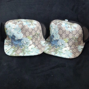Gucci keps med blå blommor - Snygg keps från Gucci med klassiskt GG-mönster och stora blå blommor. Kepsen har svart mesh på sidorna och justerbar baksida. Perfekt statement-piece för dig som vill sticka ut med exklusiv design och coolt blommönster.