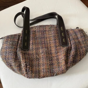 Vävd axelväska med bruna detaljer - Säljer en unik axelväska i vävt tyg med mix av beige, brun och grå toner. Väskan har breda, svarta axelband i skinnimitation med vita sömmar och coola detaljer. Perfekt för dig som gillar retrovibbar och vill sticka ut med en annorlunda väska.