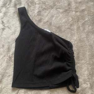 Svart oneshoulder topp från new yorker  - Svart ribbad oneshoulder topp från new yorker i storlek XS. Toppen har snörning i sidan som går att ändra och som ger en snygg rynkad effekt och croppad passform. Perfekt för dig som gillar trendiga och stilrena plagg med det lilla extra.