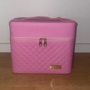Rosa quiltad sminkväska med spegel - Säljer en rosa quiltad sminkväska från Cailyn med dragkedja och praktiskt handtag. Väskan har flera utdragbara fack och en inbyggd spegel på insidan av locket. Perfekt för att organisera smink eller accessoarer. Snygg och stilren design med guldig logga framtill.