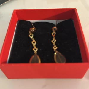 Guldiga örhängen med hjärtan och droppar - Snygga guldiga örhängen med flera små hjärtan på rad och en mörk droppformad detalj längst ner. Perfekta för dig som vill ha något unikt och romantiskt i öronen. Kommer i en röd ask.